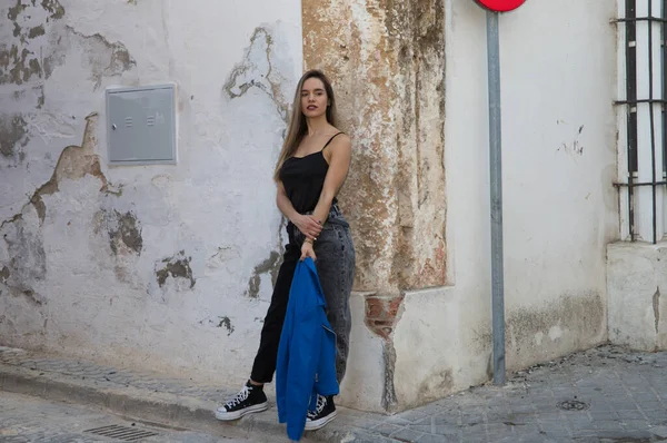 Siyah tişört ve kot pantolon giymiş genç sarışın kadın elinde mavi ceketle eski bir duvara yaslanıyor. Genç kadın farklı jestler ve vücut ifadeleri yapıyor..