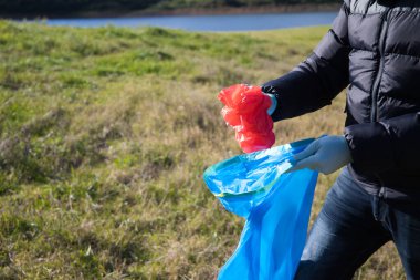 Detail of a volunteer's hand picking up a red plastic bag from the forest and putting it in a garbage bag. Concept of Earth Day and World Environment Day 5 June.