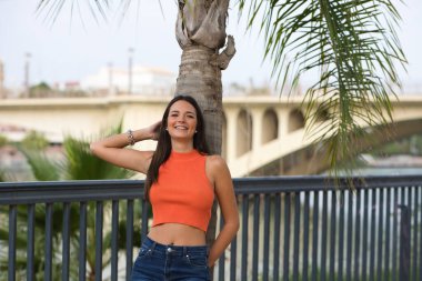 Young and beautiful woman leaning on the trunk of a palm tree. Woman is happy and relaxed and enjoying the sunny day in seville, spain. The woman is wearing jeans and orange top.