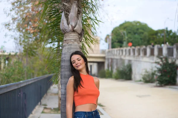 年轻美丽的女人靠在一棵棕榈树的树干上 女人很快乐 很放松 在西班牙的塞维利亚享受着阳光灿烂的一天 那个女人穿着牛仔裤和橙色上衣 — 图库照片