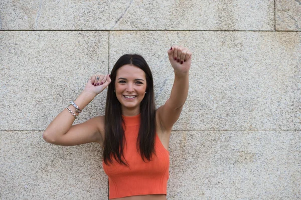 beautiful young woman raises her arms with clenched fists celebrating victory and achievements on gray background.