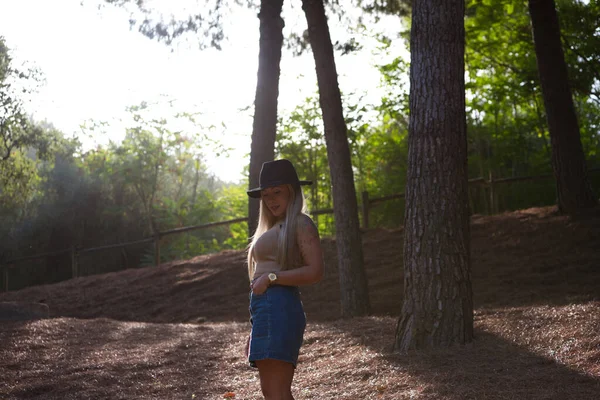 头发直的金发美女穿着棕色上衣 短短的斜纹棉裙和帽子 这个女人很快乐 在森林里的松树丛中散步 阳光从后面照进来 — 图库照片