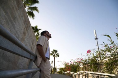 Young Latin man dressed in beige suit, white t-shirt and black boots leaning on the railing of some park stairs. Man has social problems and is stressed thinking about life's difficulties.