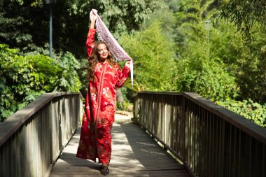 Güzel bir Faslı kadın geleneksel Fas kırmızısı, altın ve gümüş işlemeli bir elbise giyiyor. Kadın, bitkiler arasında tahta bir köprüde pembe bir eşarpla dans ediyor..
