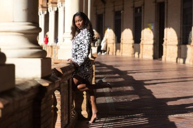 beautiful young black latin woman is in the biggest and most important square in seville, spain. She is leaning on the railing looking at the horizon of the wide square.