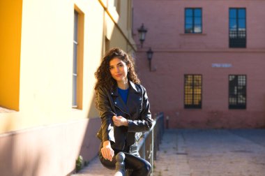 Young and beautiful Hispanic brunette woman with curly hair and black leather clothes is sitting on a railing in the street. The woman is happy and smiling.