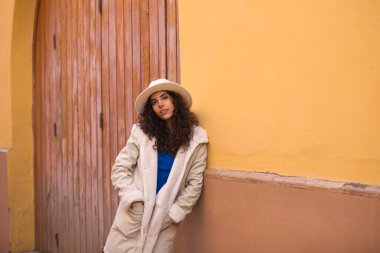 Young and beautiful Hispanic brunette woman with curly hair with hat and coat for the cold leaning on wall of a house in seville. Travel and holiday concept.