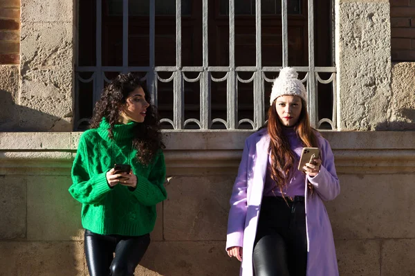 Two young and beautiful Spanish women are consulting the mobile phone in a street of a big city. The women are having fun while consulting the mobile phone.