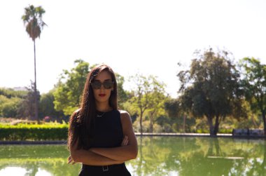 Kırmızı rujlu güzel esmer İspanyol kadın. Arka planda parkta bir göl var. Kadın zarif siyah elbiseler giymiş ve güneş gözlüğü takıyor..