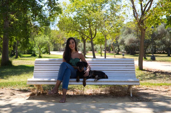 Beautiful black haired woman is sitting with her doberman puppy on a white bench in the park. The woman enjoys the company of her dog and relaxing together. Pets and animals concept.