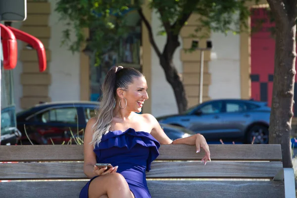 Young and beautiful blonde latina woman dressed in short dress is resting sitting on a bench in the city. The woman is consulting her mobile phone.