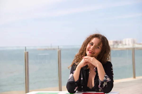 Beautiful blonde woman with curly hair and blue eyes is sitting on the promenade. In the background you can see the sea. The woman looks at camera and makes different expressions, happy, sad.