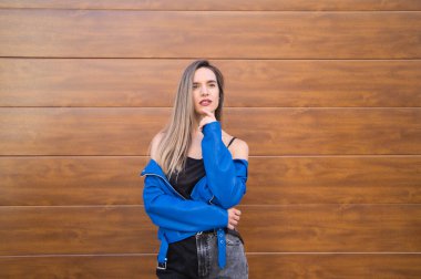 young and beautiful blonde woman dressed in black t-shirt and blue leather jacket and jeans on wooden background poses for photos doing different poses like a model. Beauty and fashion concept.