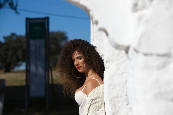 Latin woman with dark, curly hair and middle-aged dressed in jeans and white lace top is leaning on a column of a Mediterranean style house with white painted wall. The woman is on holiday.