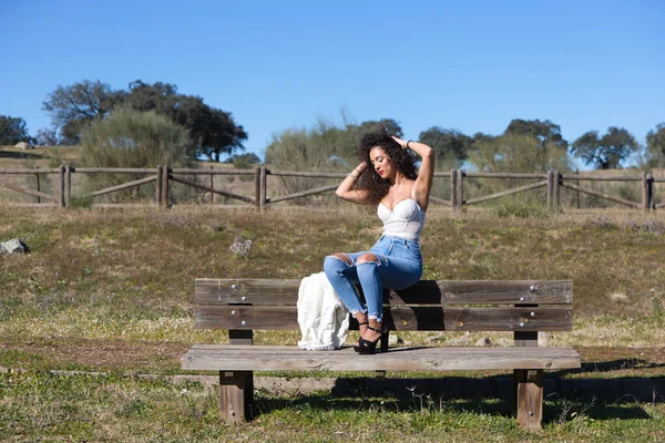 西班牙黑头发卷曲的女人坐在通向森林的小路上的长椅上 她穿着牛仔裤和白色上衣 享受着阳光灿烂的日子 — 图库照片