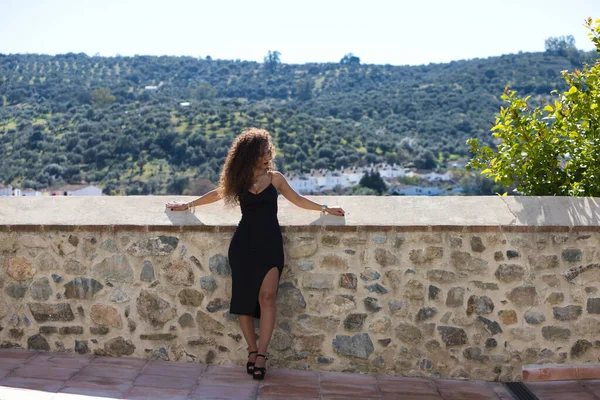 Güzel, siyah saçlı, kıvırcık saçlı ve zarif elbiseli bir kadın aşağıya bakıyor, duvara yaslanıyor. Arka planda dağı ve dağdaki ağaçları görebilirsiniz. Kadın üzgün..