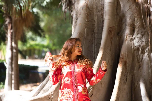 A beautiful Moroccan woman is wearing a traditional Moroccan red dress with gold and silver embroidery. The woman is in a park next to a tree trunk. She enjoys the trip to the city.