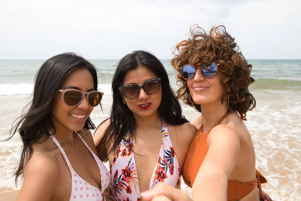 stock image Three beautiful women taking a selfie on the beach with the sea in the background. The women are on holiday and are smiling for the photo, wearing sunglasses. Holiday and travel concept.