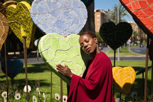 Young and beautiful Afro American woman hugging hearts of different sizes and colors in a park. Valentine's Day