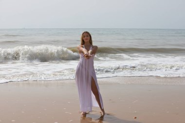 Beyaz elbiseli güzel sarışın kadın elinde bir midye ya da deniz kabuğu tutuyor ve onu ön tarafa gösteriyor. Kadın mutlu ve eğleniyor. Arka planda mavi deniz.