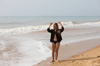 Genç ve güzel sarışın kadın siyah gömlek ve siyah bikini giyip sahilde yürüyor ve farklı ifadeler ve duruşlar sergiliyor. Arka planda deniz ve gökyüzü.