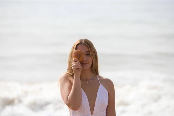 Pretty young blonde woman in white dress holds a clam or shell in her hand and covers one eye while smiling. The woman is happy and having fun. In the background the blue sky and sea.