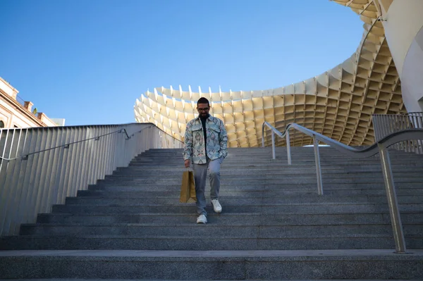 stock image Handsome young man with beard comes down stairs with shopping bag in hand. Man is dressed casually and in modern clothes. Shopping and holiday concept.