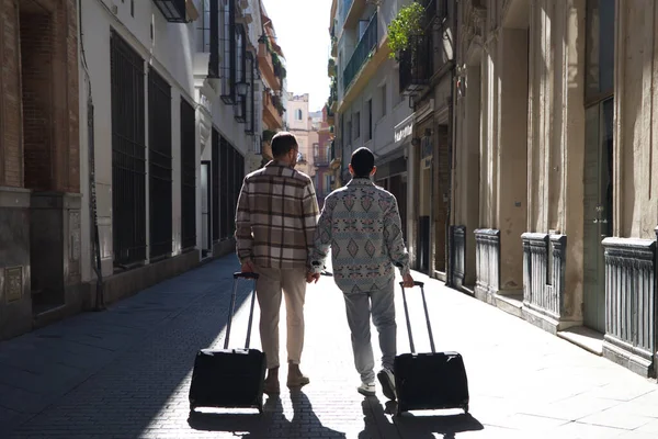 若いゲイの男性カップルがスーツケースを持って通りを歩いている 二人は旅行に行く 写真は後ろから撮影されています 休暇と旅行のコンセプト — ストック写真
