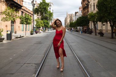 Kırmızı ipek elbiseli güzel bir kadın Seville 'de bir caddede tramvay raylarının arasında duruyor. Model fotoğraf çekimi için poz veriyor. Moda ve güzellik konsepti.