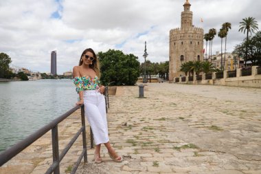 Young, pretty blonde woman with sunglasses leaning on the railing of the river. The woman is happy and smiling. In the background you can see the river in seville.
