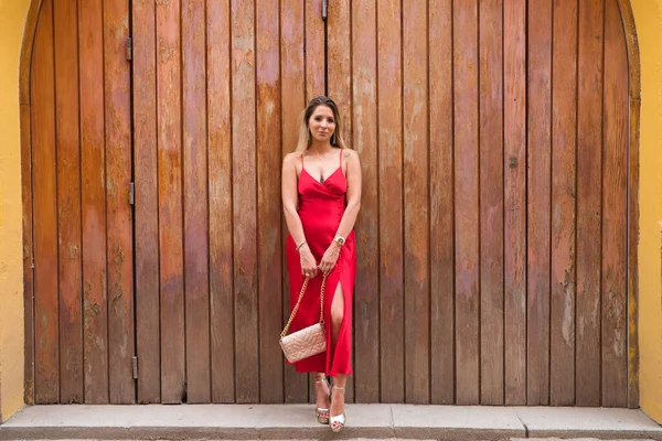 Young beautiful woman in red silk dress and pink handbag leaning against a wooden door poses for photo. Concept expressions and gestures