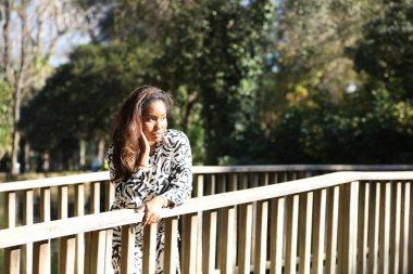 beautiful young black latin woman leaning on the railing of a wooden bridge in a park in seville looking at the camera with her face in her hands.