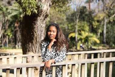 beautiful young black latin woman leaning on the railing of a wooden bridge in a park in seville looking at the camera with her face in her hands.