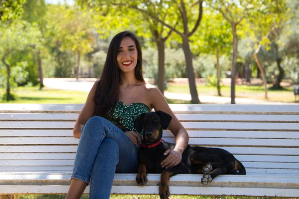 Beautiful black haired woman is sitting with her doberman puppy on a white bench in the park. The woman enjoys the company of her dog and relaxing together. Pets and animals concept.