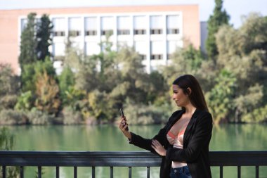 Young beautiful woman looking at her mobile phone leaning on the railing of the river in Seville, Spain. In the background you can see the river and part of the city.
