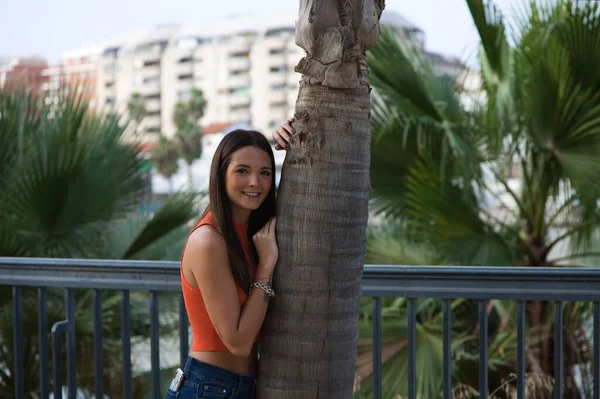 若くて美しい女性はヤシの木の幹にもたれています 女性は幸せとリラックスしていて スペインのセビリアで晴れた日を楽しんでいます 女性はジーンズとオレンジの上を着ている — ストック写真