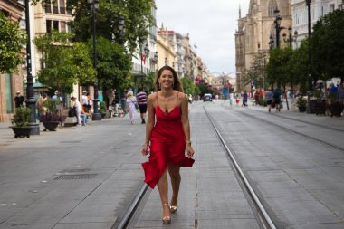Büyük bir şehirde tramvay rayları arasında koşan kırmızı ipek elbiseli güzel bir kadın. Manken tek eliyle elbisesini tutuyor. Moda ve güzellik konsepti.