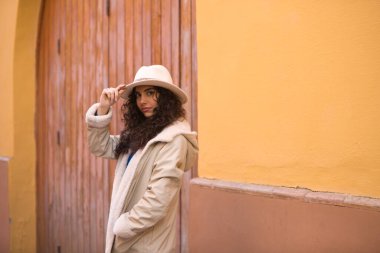 Young and beautiful Hispanic brunette woman with curly hair with hat and coat for the cold leaning on wall of a house in seville. Travel and holiday concept.