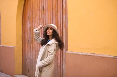 Young and beautiful Hispanic brunette woman with curly hair wearing a hat and coat for the cold walking in the city of seville while making different expressions and having fun.