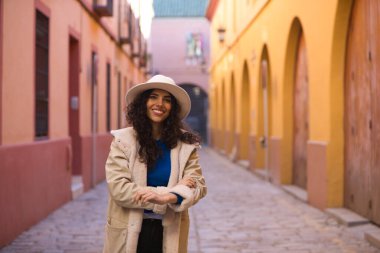 Young and beautiful Hispanic brunette woman with curly hair wearing a hat and coat for the cold walking in the city of seville while making different expressions and having fun.