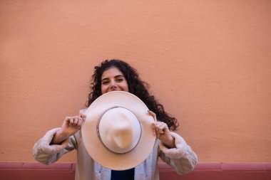 Young and beautiful Hispanic brunette woman with curly hair covers her face with her hat and exposes her eyes. The woman plays at hiding behind the hat making different funny expressions.