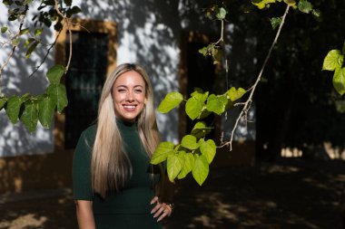 Yeşil elbiseli güzel sarışın kadın bir ışık huzmesinin geçtiği ağacın dallarının yanında duruyor. Doğayla temas.