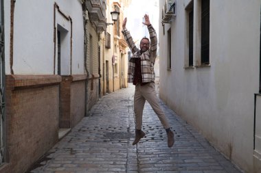 Sakallı ve gözlüklü yakışıklı genç bir adam dar bir sokakta sevinçten zıplıyor. Mutluluk ve neşe kavramı.