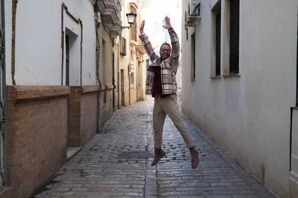 Sakallı ve gözlüklü yakışıklı genç bir adam dar bir sokakta sevinçten zıplıyor. Mutluluk ve neşe kavramı.