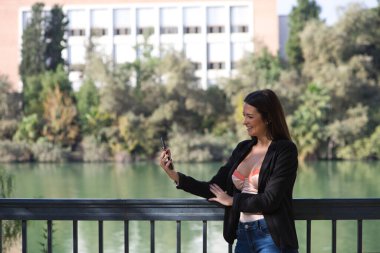 Young beautiful woman looking at her mobile phone leaning on the railing of the river in Seville, Spain. In the background you can see the river and part of the city.