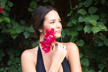 Young beautiful brunette typical Spanish woman dressed in black suit and bouquet of pink flowers near her face. The woman is in the park surrounded by greenery. The woman is happy.