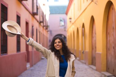 Young and beautiful Hispanic brunette woman with curly hair holds up a hat with her hand while making different expressions and having fun on her city trip in Europe.