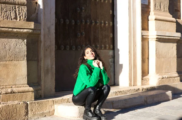 巻き毛と緑のウールのコートを着たかなり若いブルネットの女性は スペインのセビリアの建物の入り口の一歩に座っています 女性は幸せと街で彼女の休日を楽しんでいます — ストック写真