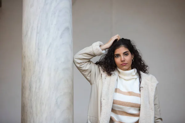 stock image Young and beautiful brunette woman with curly hair and a woollen coat for the winter, leaning on a marble column while making different facial and body expressions.