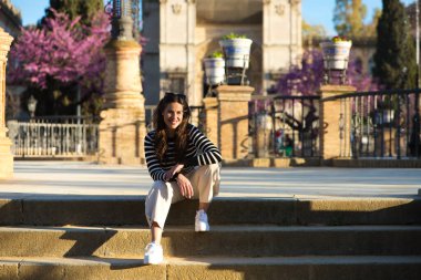 Uzun kahverengi saçlı, siyah beyaz tişörtlü ve beyaz pantolonlu güzel bir kadın Seville 'deki bir parkın merdivenlerinde oturuyor..